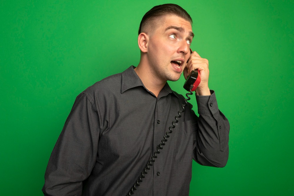 young handsome man grey shirt talking vintage phone looking aside confused standing green wall